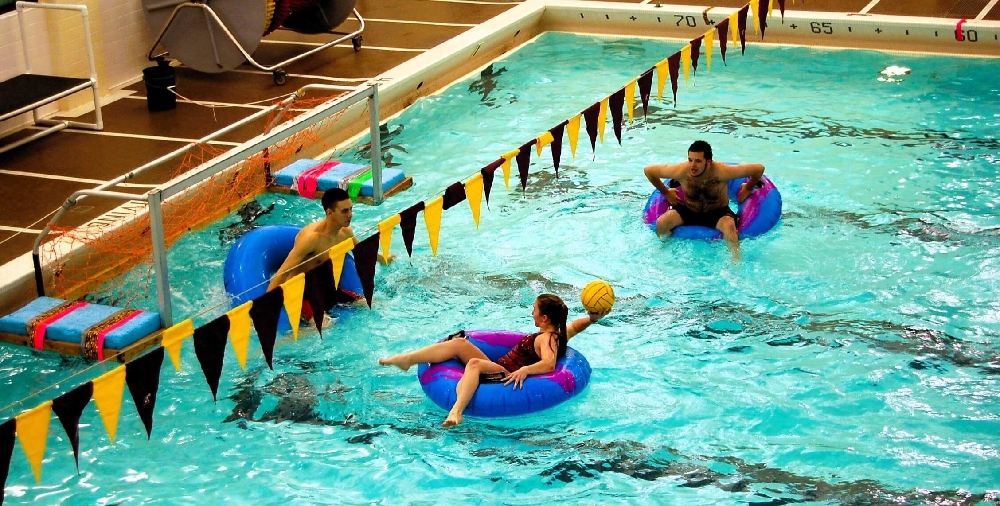 Students playing inner tube water polo in a campus pool, floating on inflatable rings while tossing a yellow ball. Central Michigan University Inner Tube Water Polo.